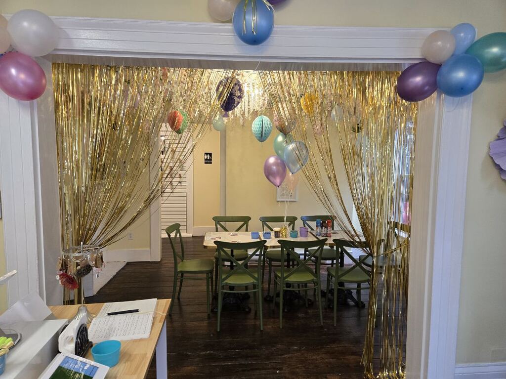 The party room seen through a gold fringe curtain, with balloons over the doorway and a long table set for a group.