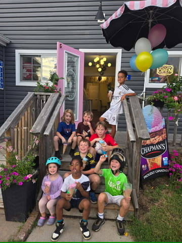 A birthday group on the front steps of Ice Cream on Elm, cones in hand, under the striped awning and balloons.