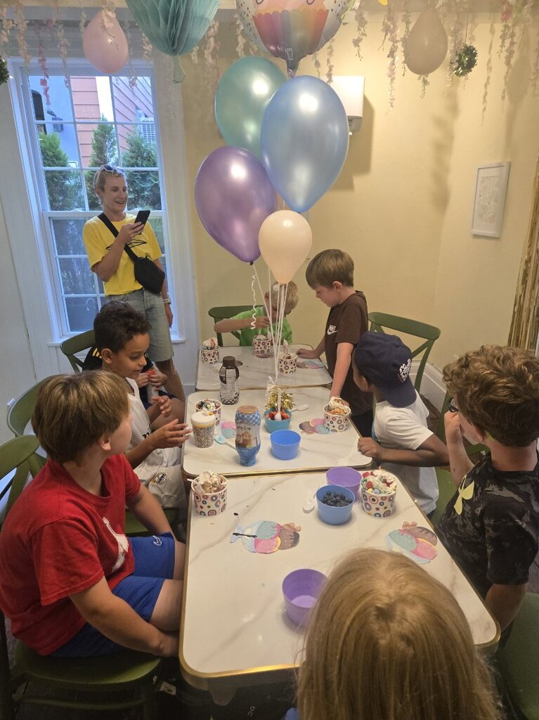 A group of kids around the party table digging into sundae cups, with a bunch of pastel balloons tied to the middle of the table.