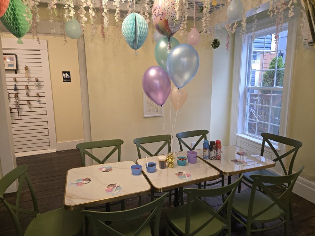 Tables set for a birthday with sundae cups, sprinkles and toppings, under pastel balloons and paper lanterns.